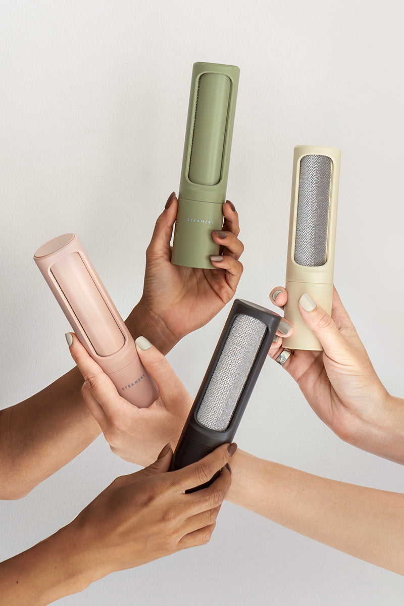Vier Hände zeigen die vertikalen, zylindrischen, wiederverwendbaren Steamery-Fusselrollen in Pastellrosa, Grün, Beige und Anthrazit. Jede Bürste verfügt über eine metallisch strukturierte Reinigungsoberfläche und ein minimalistisches Design vor einem cremefarbenen Hintergrund.