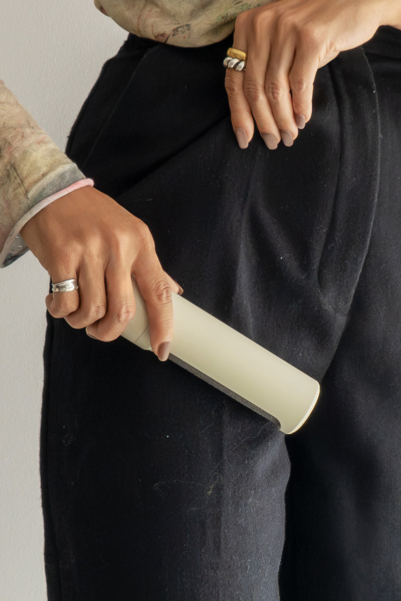 Eine Person benutzt einen beigen, wiederverwendbaren Steamery Flusenentferner auf einer schwarzen Hose, um Fussel oder Staub zu entfernen. Ihre silberumrandete Hand hält die Hose vor einem unifarbenen Hintergrund fest, wodurch sowohl die Hände als auch der Reinigungsvorgang hervorgehoben werden.