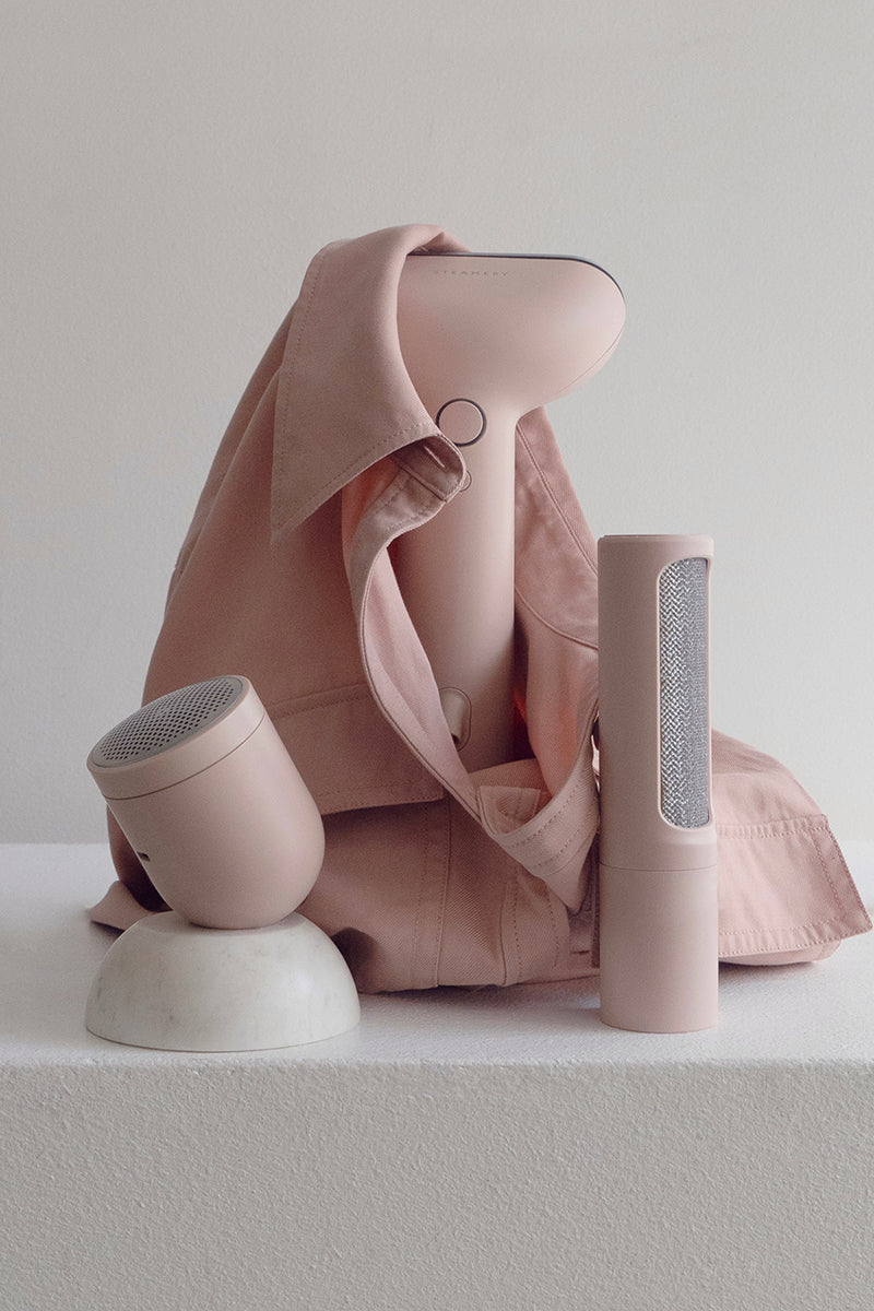 Ein blassrosa Steamery Pilo 2 Handbedampfungsgerät für Kleidung steht aufrecht mit einem passenden Hemd. Daneben sind zwei weitere blassrosa Steamery-Produkte - ein Fusselentferner und ein Stoffrasierer - auf einer weißen, strukturierten Fläche ausgestellt.