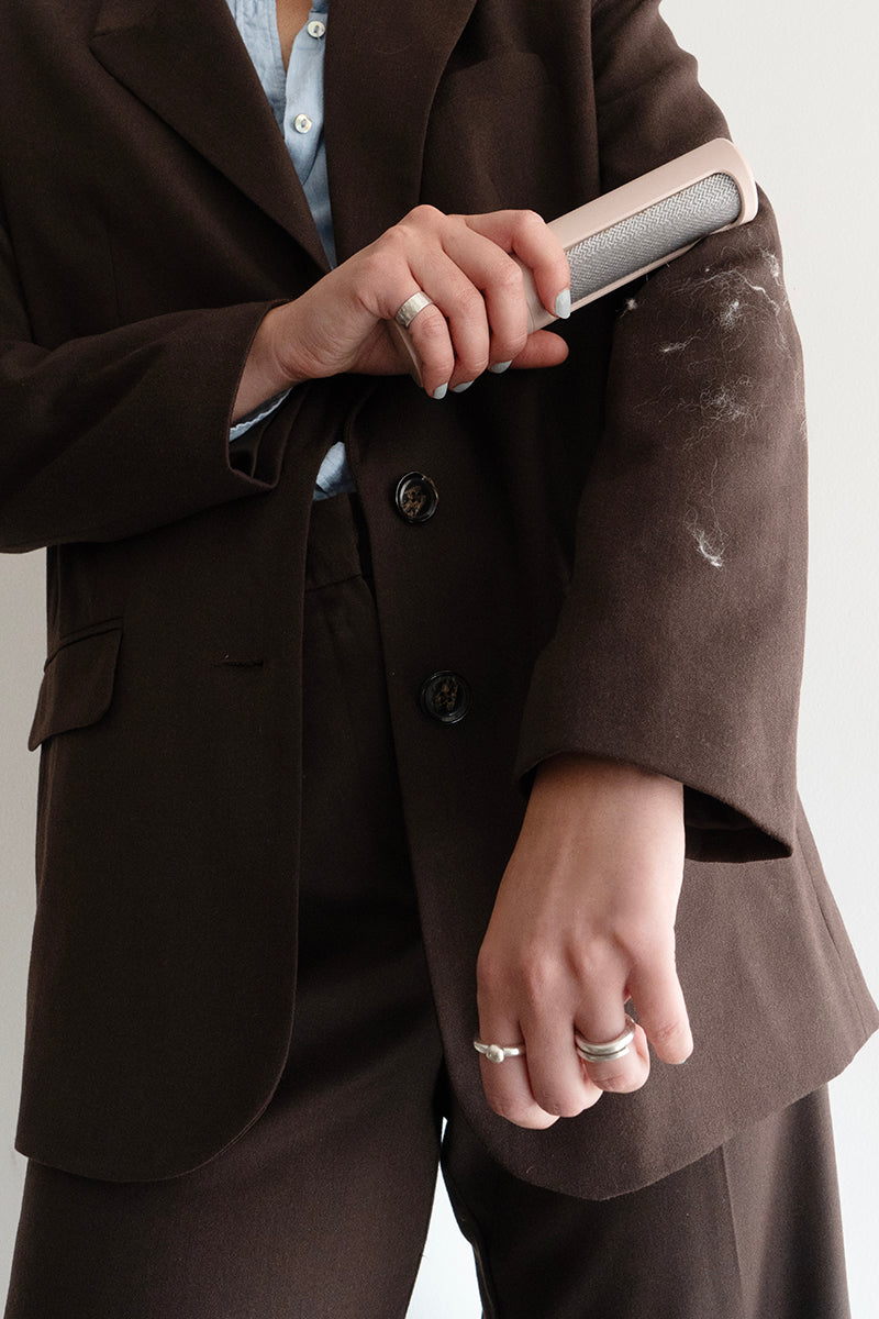 Eine Person in einem braunen Anzug benutzt einen wiederverwendbaren Flusenentferner, um weiße Flusen von ihrem Ärmel zu entfernen, wobei ein hellblaues Hemd darunter und silberne Ringe an ihren Fingern zum Vorschein kommen.