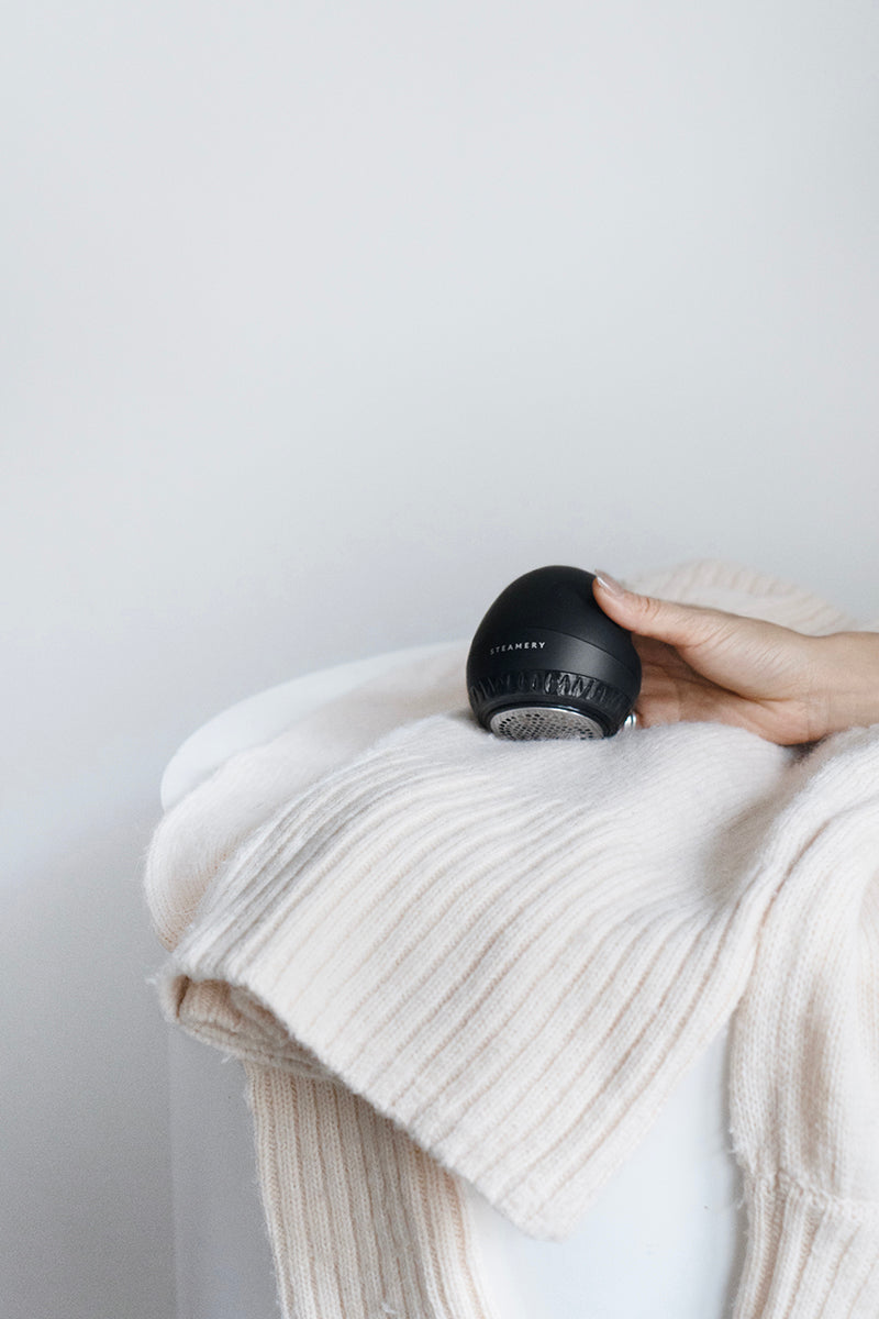 Eine Hand hält einen schlanken, schwarzen Steamery-Fusselrasierer über einem weichen, cremefarbenen Strickpullover, der auf einer weißen Fläche mit einem minimalistischen hellen Hintergrund drapiert ist.