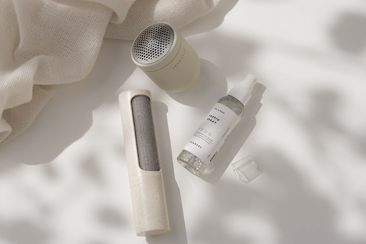 Drei minimalistische Dampferzeugnisse auf einer weißen Fläche: ein kleiner runder Lautsprecher, eine klare "Spring"-Sprühflasche mit weißem Deckel und ein wiederverwendbarer Fusselentferner. Außerdem sind weiche Blattschatten und ein strukturierter beiger Stoff zu sehen.