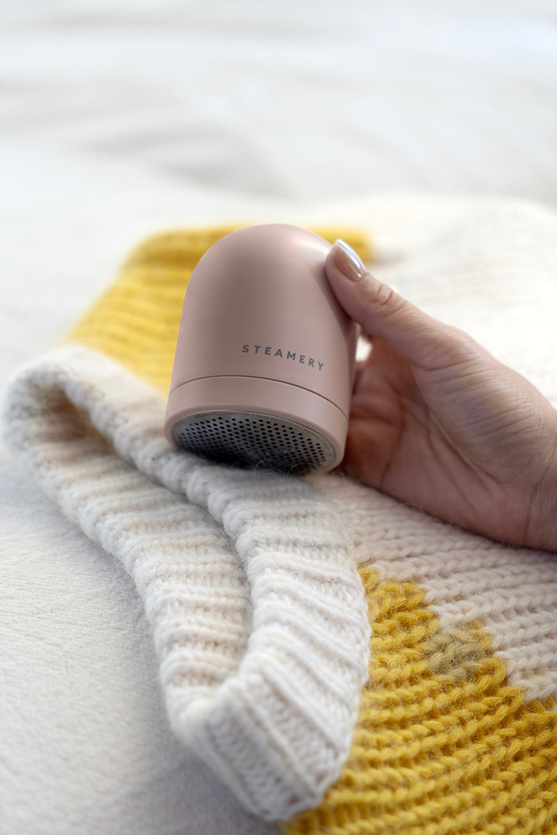 Eine Hand hält einen rosafarbenen Steamery Pilo 2 Stoffrasierer über einen weiß-gelben Strickpullover und zeigt damit die sanfte Entfernung von Stoffpillen an, um das Aussehen des Pullovers zu erhalten.