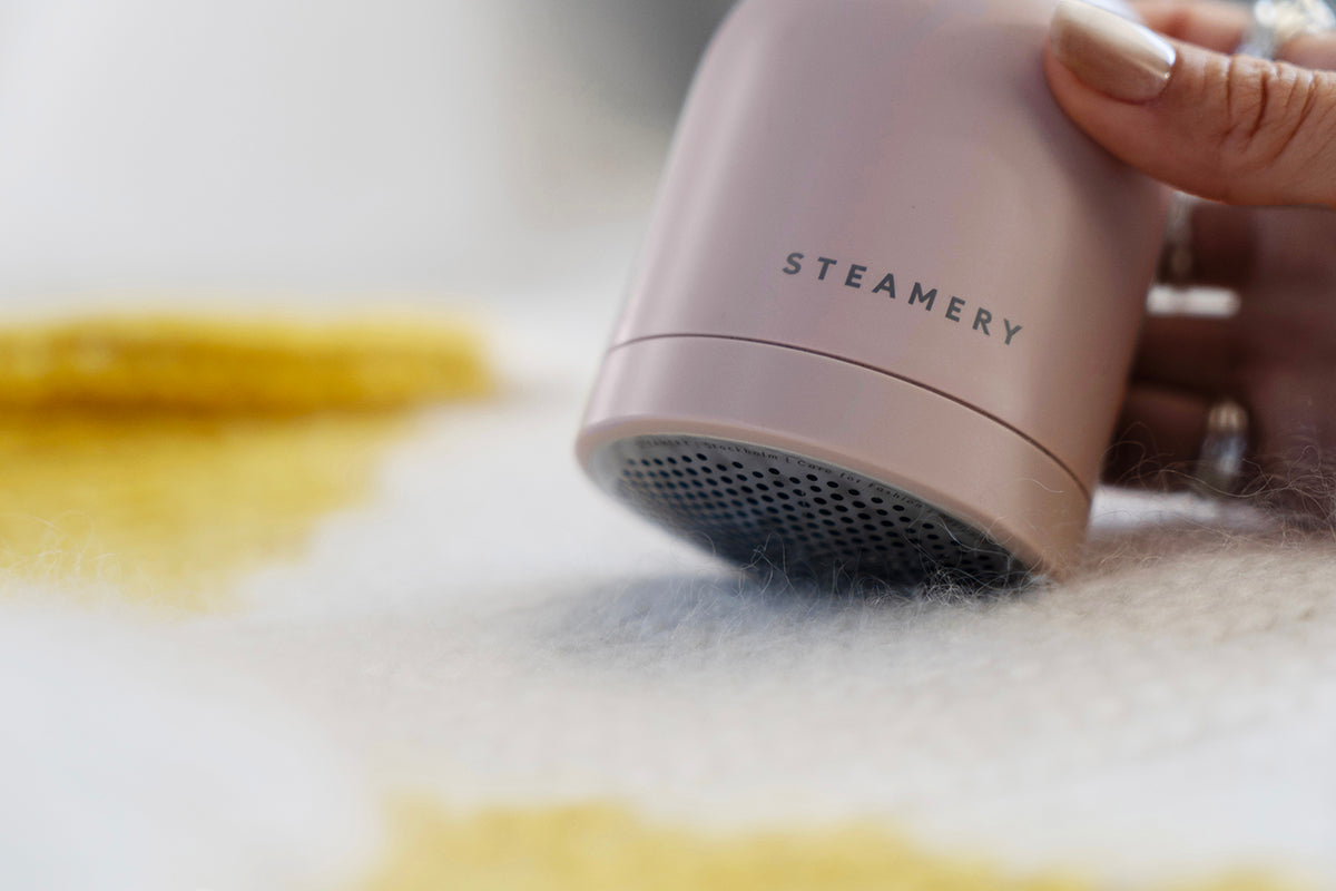 Eine Hand hält einen rosafarbenen Steamery Pilo 2-Rasierapparat und lässt ihn über weißes und gelbes Gewebe gleiten, um Fusseln zu entfernen. Die Nahaufnahme zeigt die Metallgitteroberfläche des Rasierers und die sauber manikürten Nägel der Person.