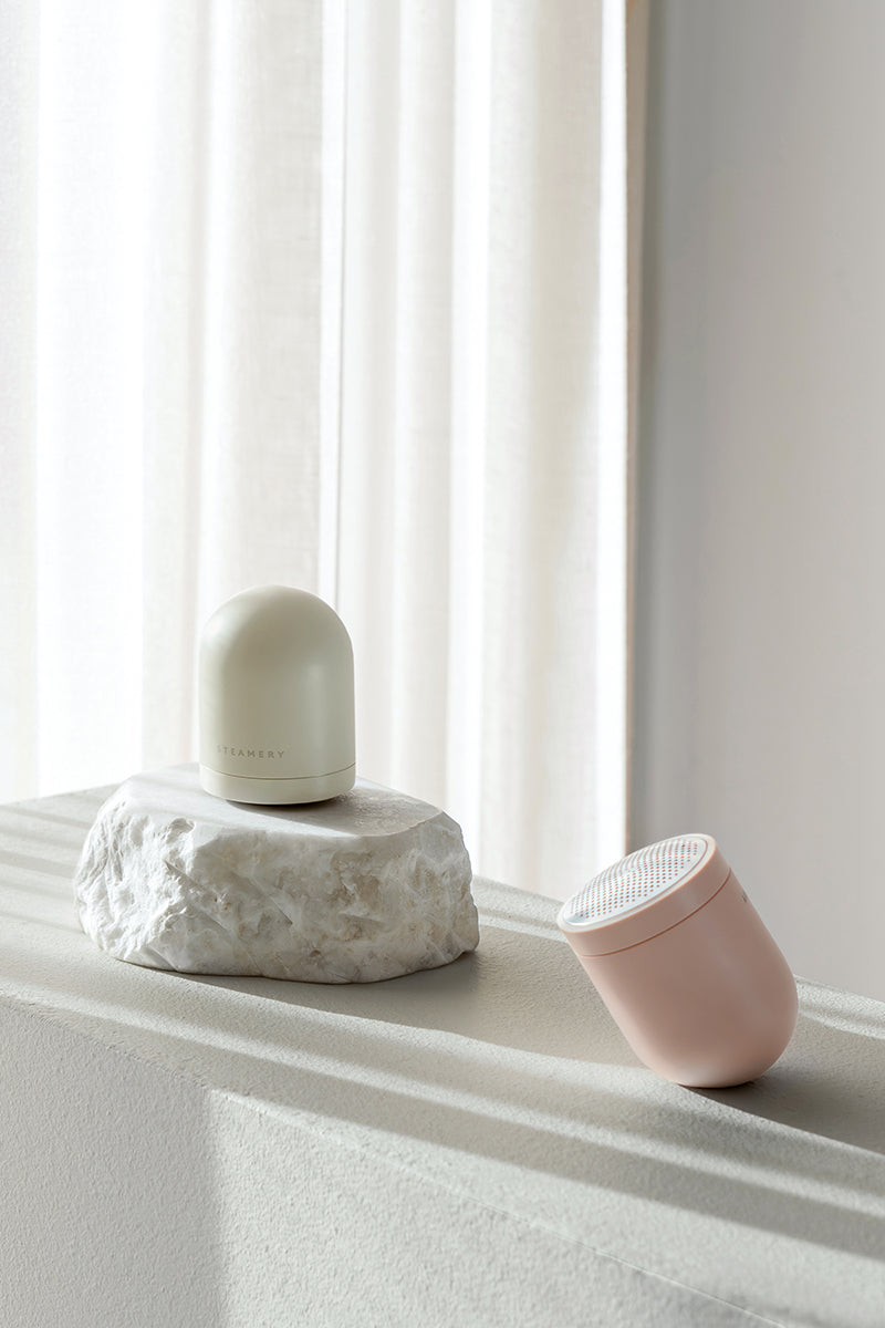 Zwei minimalistische Steamery-Hautpflegegeräte ruhen auf einer hellen Fläche: Der weiße, kuppelförmige Pilo 2 Fusselrasierer liegt auf einem rauen Stein, während ein blassrosa Gerät mit Borsten daneben steht. Sanftes Sonnenlicht fällt durch die transparenten Vorhänge im Hintergrund.