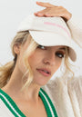A woman with wavy blonde hair in a loose ponytail wears the cream Kappe Princess Goes Hollywood by Princess Goes Hollywood, adjusting its Schirm. She pairs it with a white sweater featuring green-striped trim over a white top, gazing at the camera.